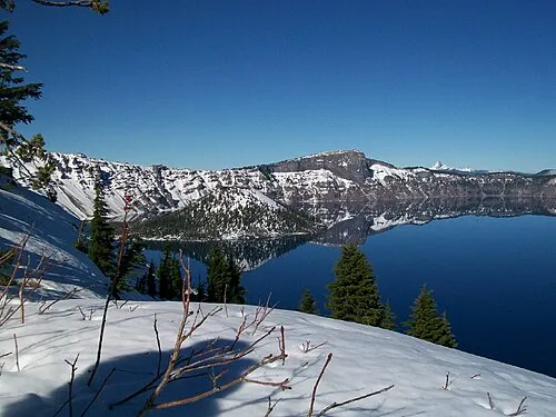Deep blue Crater Lake in early spring with the cinder cone of Wizard Island rising near the snow-edged west rim.