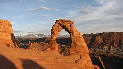 Delicate Arch, a freestanding sandstone arch about 52 feet tall, lit orange at sunset with the La Sal Mountains behind it.