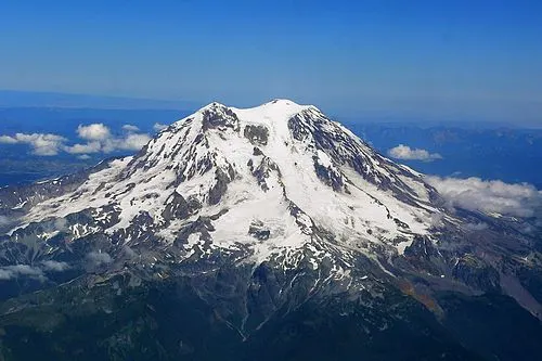 Snow-covered west face of Mount Rainier rising above forested ridges, the south-side approach that holds the Paradise meadows.