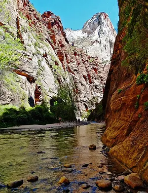Hikers wade the Virgin River where the Narrows slot canyon walls close to a narrow gap and rise hundreds of feet on both sides.
