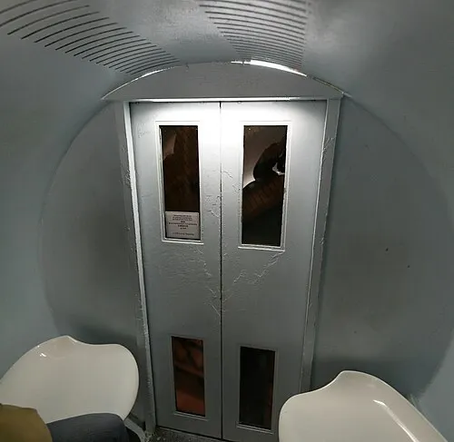 Inside a five-seat tram capsule of the Gateway Arch, a small curved white pod with bench seats and a single round window.